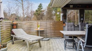 En udendørs patio med et træbord og stole, en liggestol og et lille bord med en vase blomster.