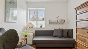 En moderne stue med en grå sofa, et træskab, et lille bord med en lampe og et vindue med udsigt til træer.