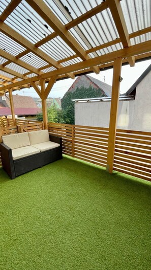 Un espace extérieur abrité, doté d’une pelouse artificielle verte, d’un canapé en osier et d’une clôture en bois.