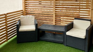 Un patio couvert comprenant deux fauteuils en osier et une petite table, avec un plafond à lattes en bois et un revêtement de sol en gazon synthétique vert.