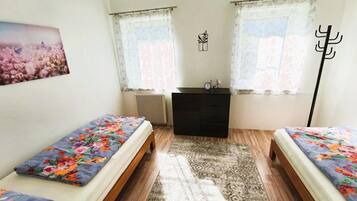 A bedroom with two beds, a dresser, a radiator, and two windows with curtains.