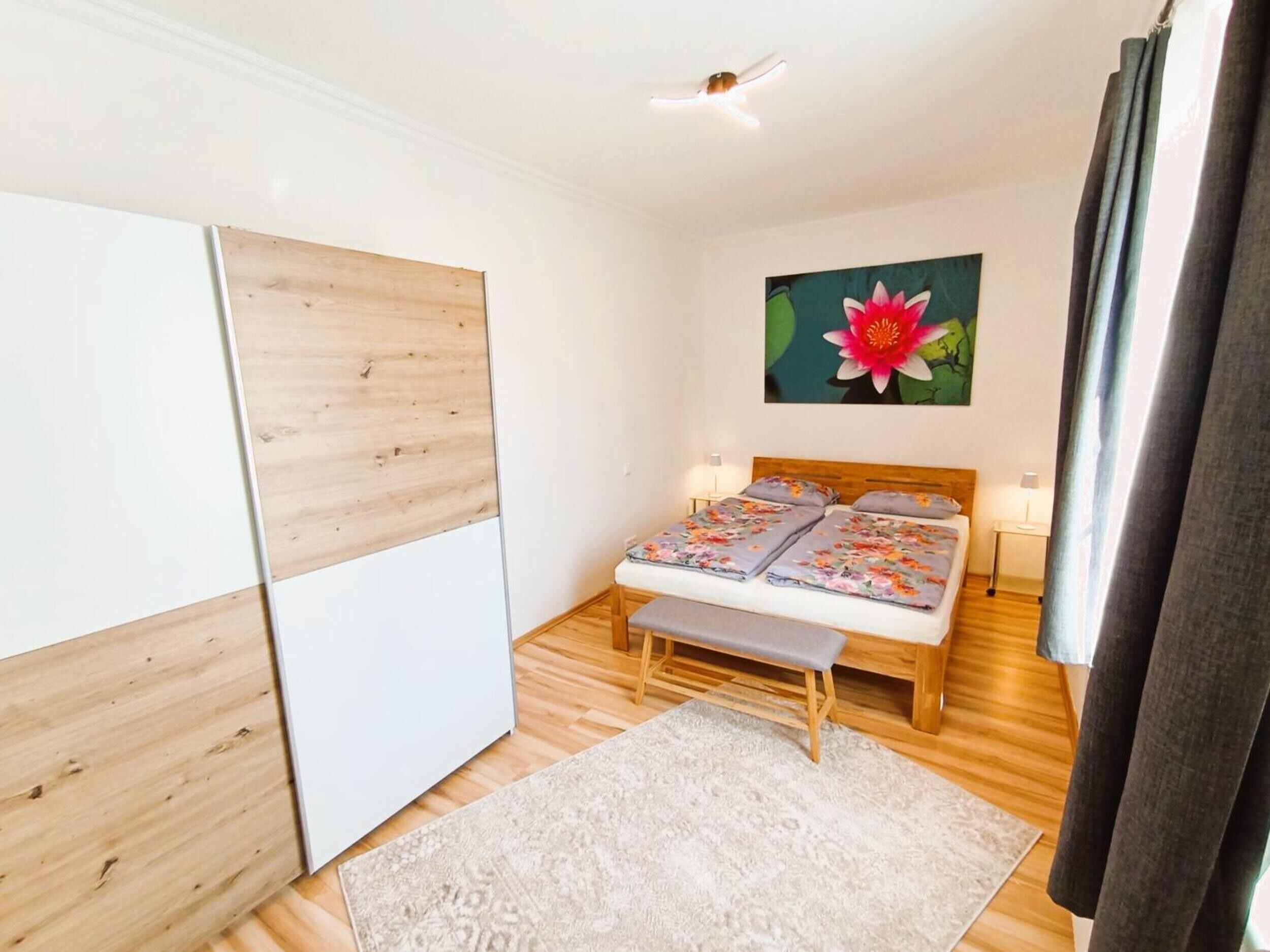 Une chambre à coucher avec un lit, une table de chevet, une armoire en bois, un tableau représentant une fleur et une fenêtre avec des rideaux.