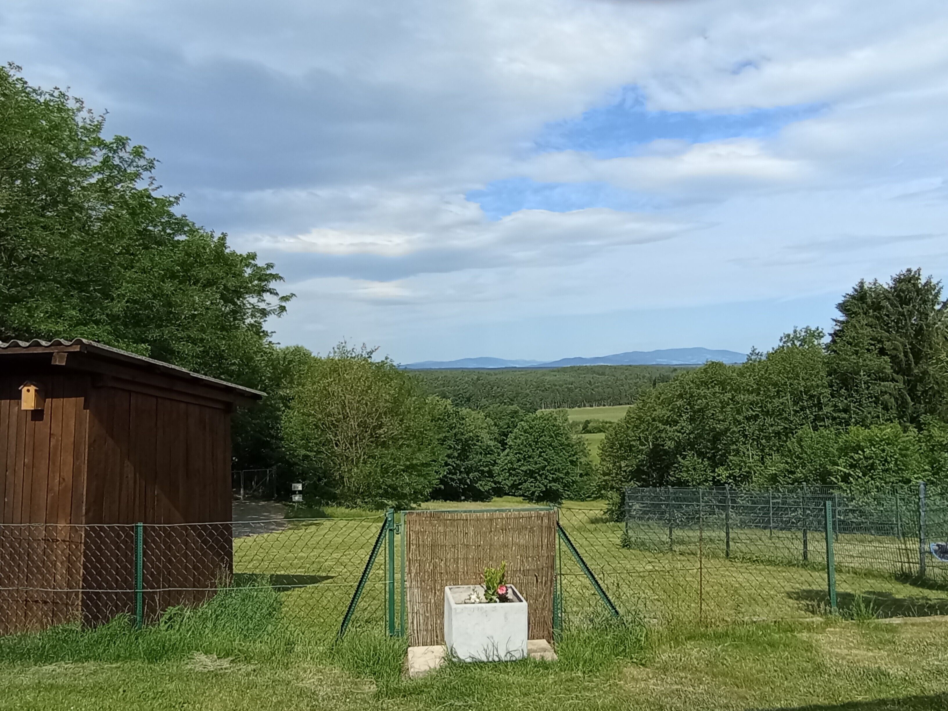 A rural landscape with a wooden shed, a fenced area with a planter box, and a view of distant hills.