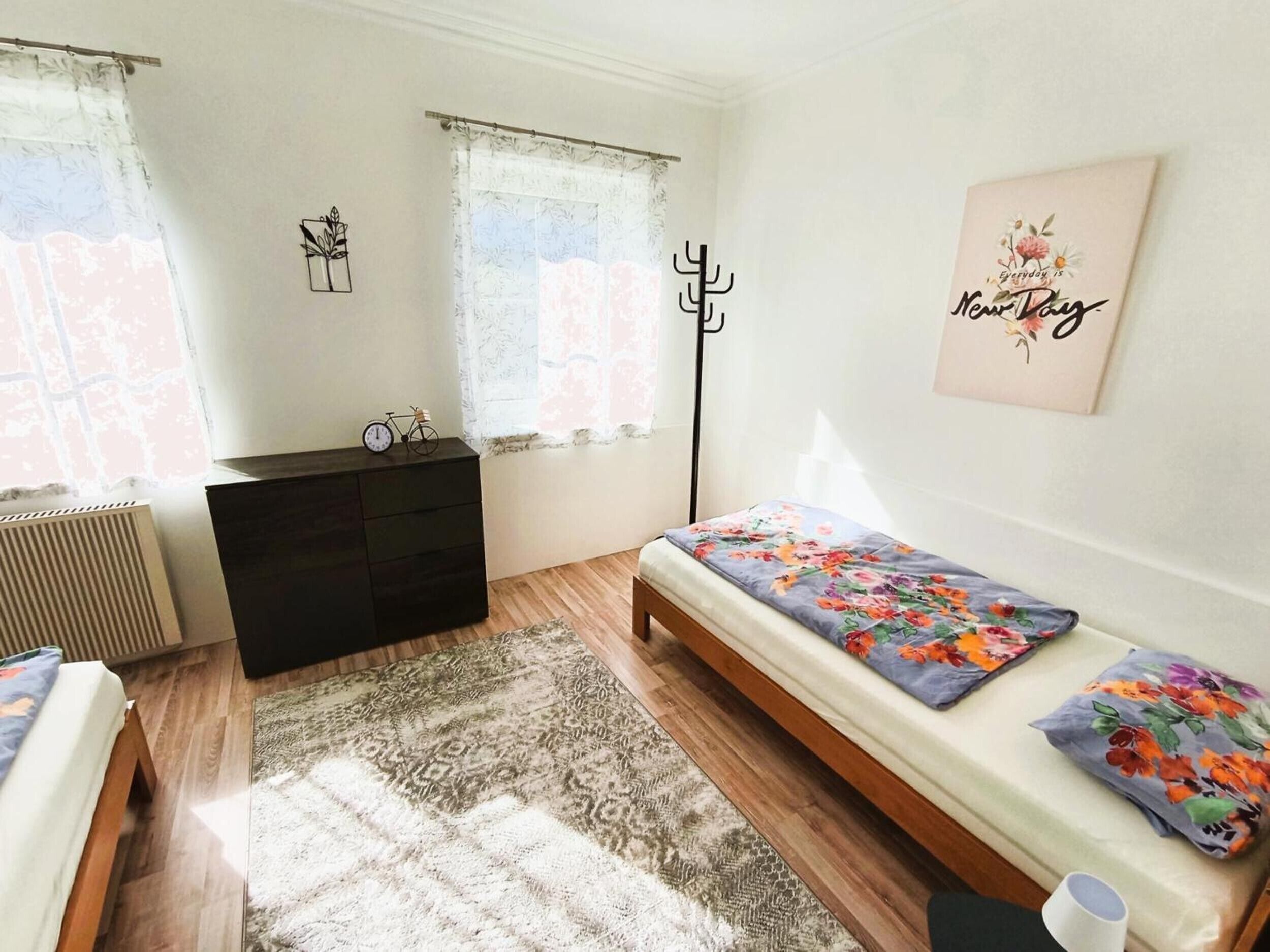 Une chambre à coucher comprenant un lit, une commode, un radiateur, une fenêtre avec des rideaux, une horloge murale et une affiche motivante.