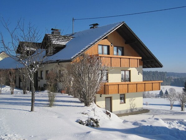 Una casa di legno a due piani con un tetto spiovente, circondata da terreno e alberi imbiancati dalla neve.