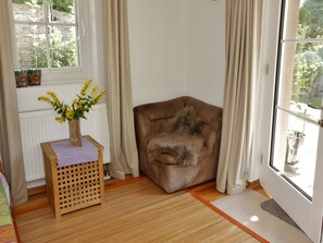 Ein Zimmer mit Holzboden, einem braunen Sessel, einem kleinen Holztisch mit einer Vase gelber Blumen und einem Fenster mit Vorhängen.