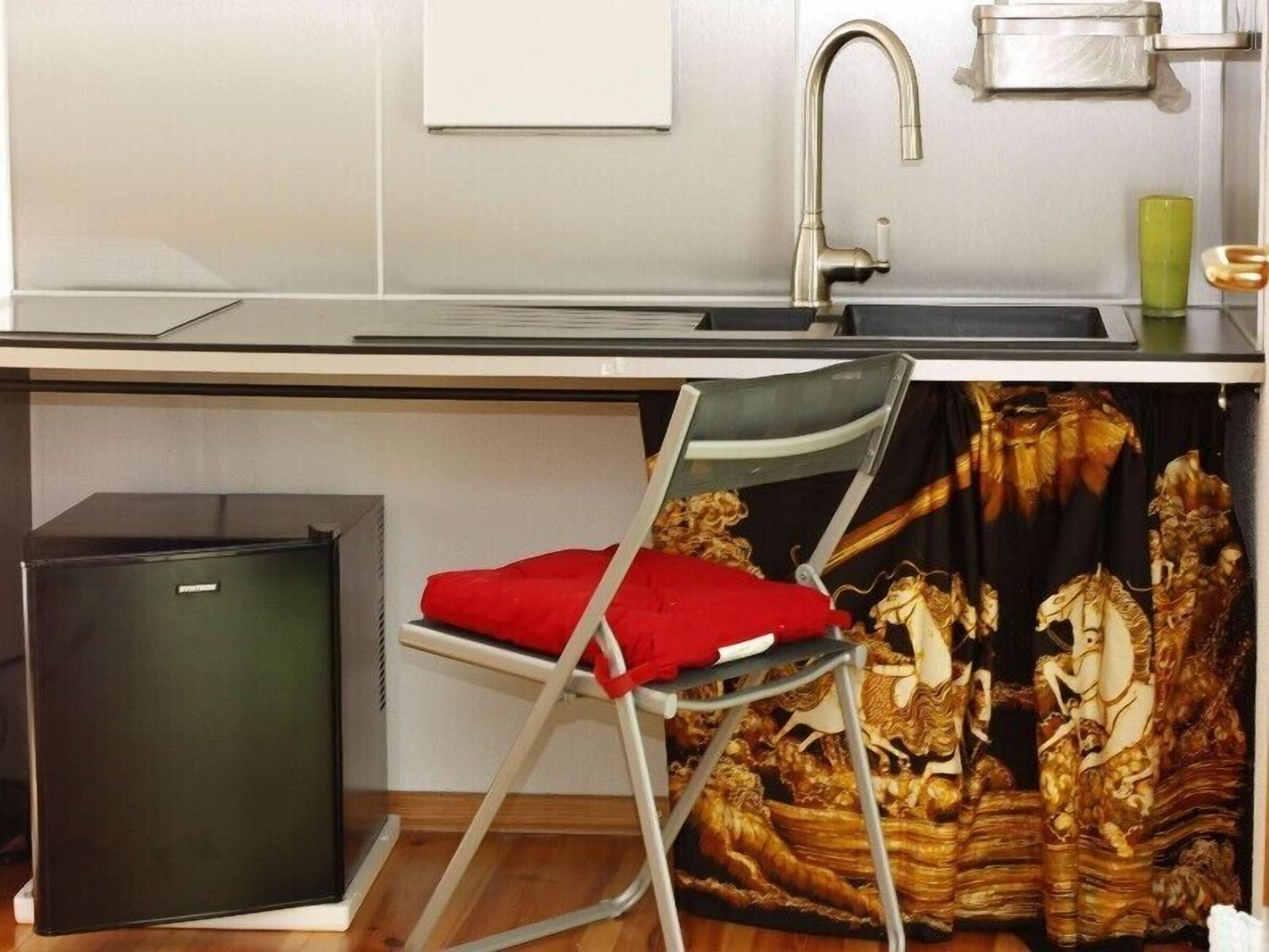 A compact kitchen with a sink, a small refrigerator, and a chair with a red cushion.