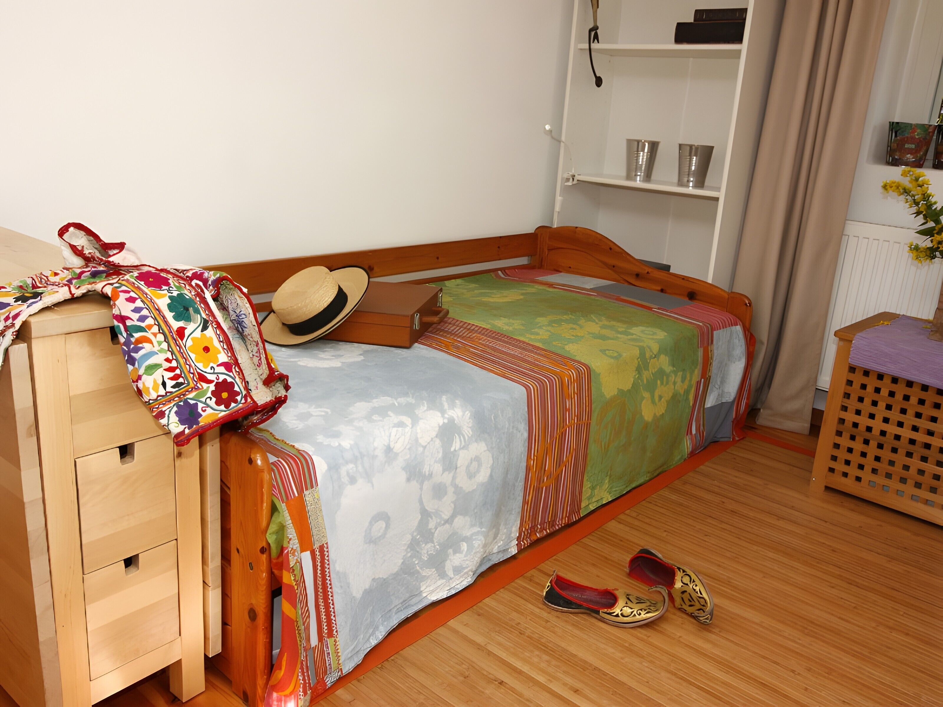 Un dormitorio con una cama de madera, un pequeño aparador de madera, un sombrero y un par de zapatos en el suelo.