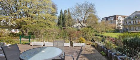 Une terrasse extérieure avec une table ronde et des chaises, entourée de verdure et offrant une vue sur des immeubles d’habitation.