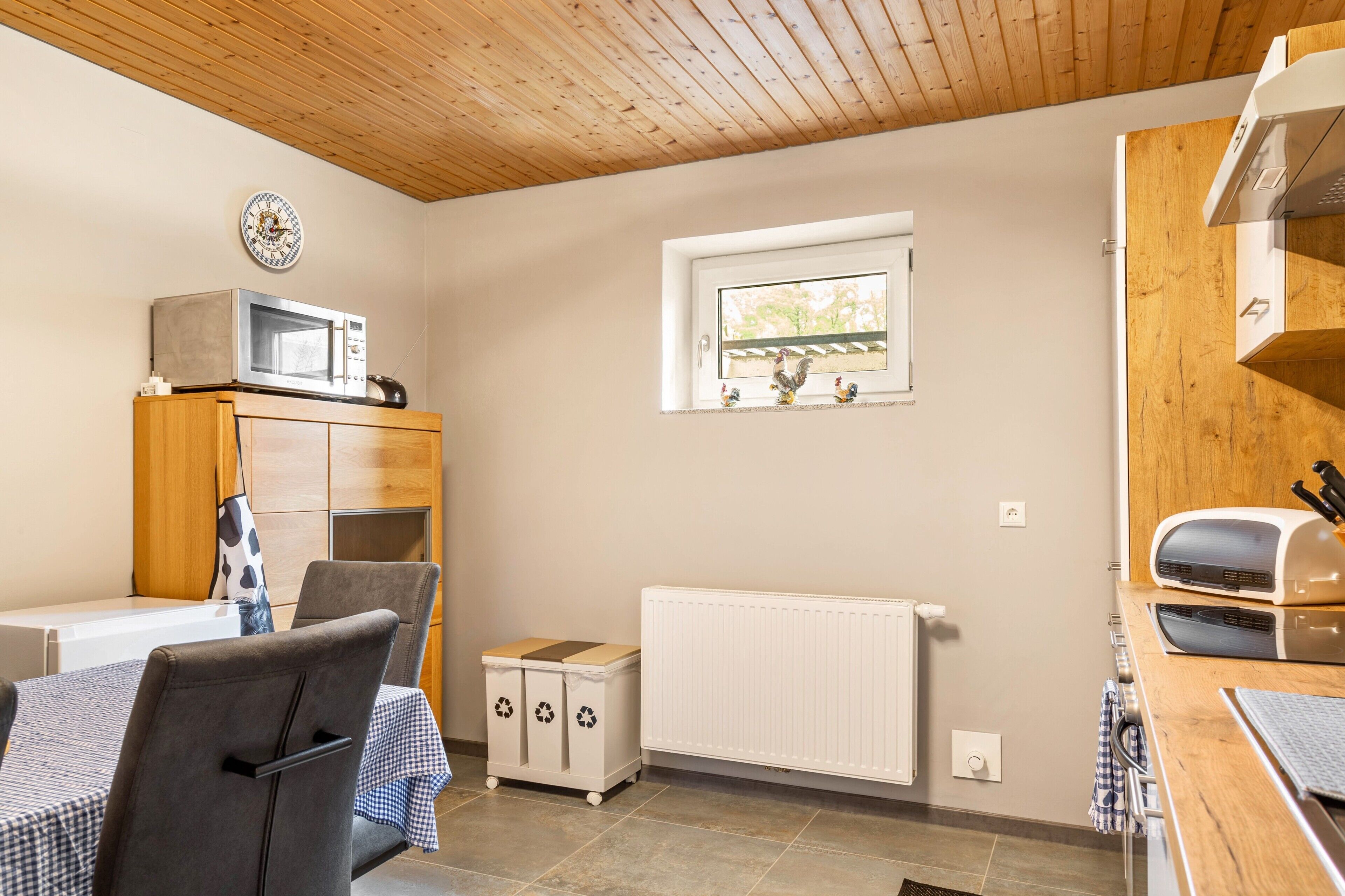 A compact kitchen with a microwave, a small window, a radiator, and a wooden ceiling.