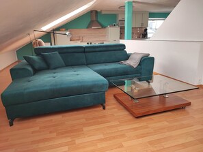 A teal sectional sofa, a glass coffee table, and a kitchen in the background.