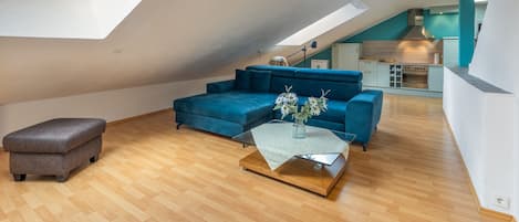 A modern loft with a sloped ceiling, a blue sofa, a wooden coffee table, and a kitchen area.