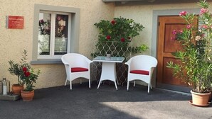 A small outdoor seating area with two white wicker chairs and a table, surrounded by potted plants and a wooden door.