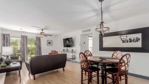 A modern living room with a black sofa, wooden dining table with chairs, a ceiling fan, and a TV mounted on the wall. - Fashion Square Townhouse | 4 Heated Pools (Scottsdale)