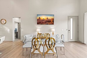 A dining area with a glass table, white chairs, and a desert sunset painting on the wall.