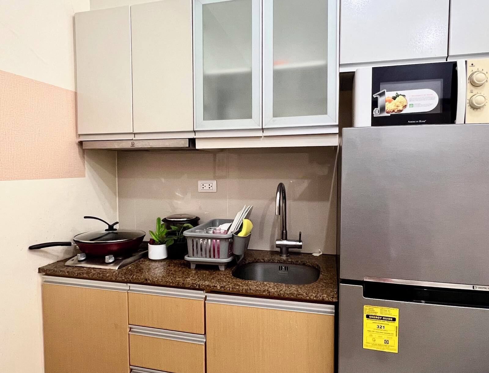 A compact kitchen with a stainless steel refrigerator, a microwave, and a sink with a faucet. There are cabinets above the countertop, and a small potted plant on the counter.