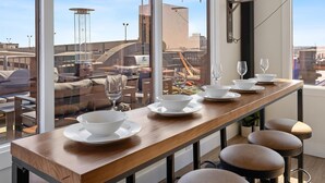 A dining area with a long wooden table, white bowls, and wine glasses, overlooking a cityscape with buildings and construction. - Penthouse of all Penthouses (Indianapolis)