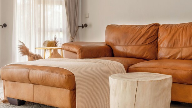 En brun lædersofa med tilhørende skammel, et træsidebord og en rund træstol i en moderne stue med lette gardiner og en lysegrå løber.