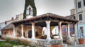 Un edificio de piedra tradicional con tejado de tejas, una torre de campanas y un patio cubierto con columnas de piedra y plantas en macetas.