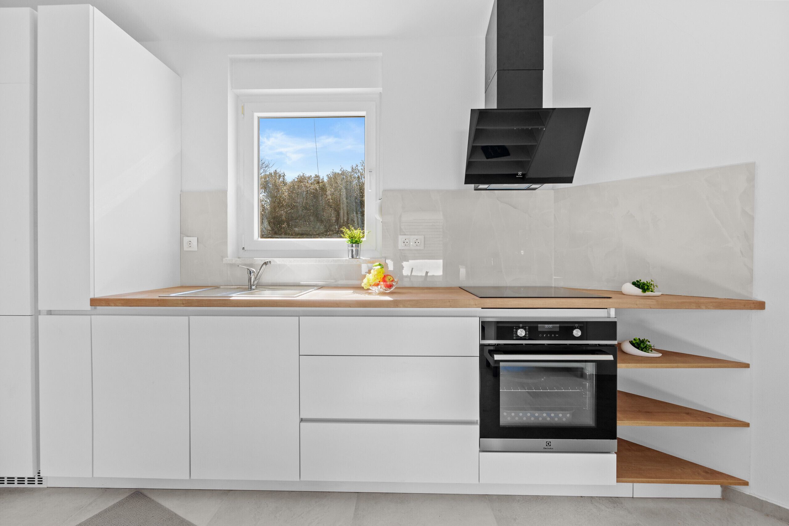A modern kitchen with white cabinetry, a wooden countertop, a built-in oven, and a window with a view of trees.
