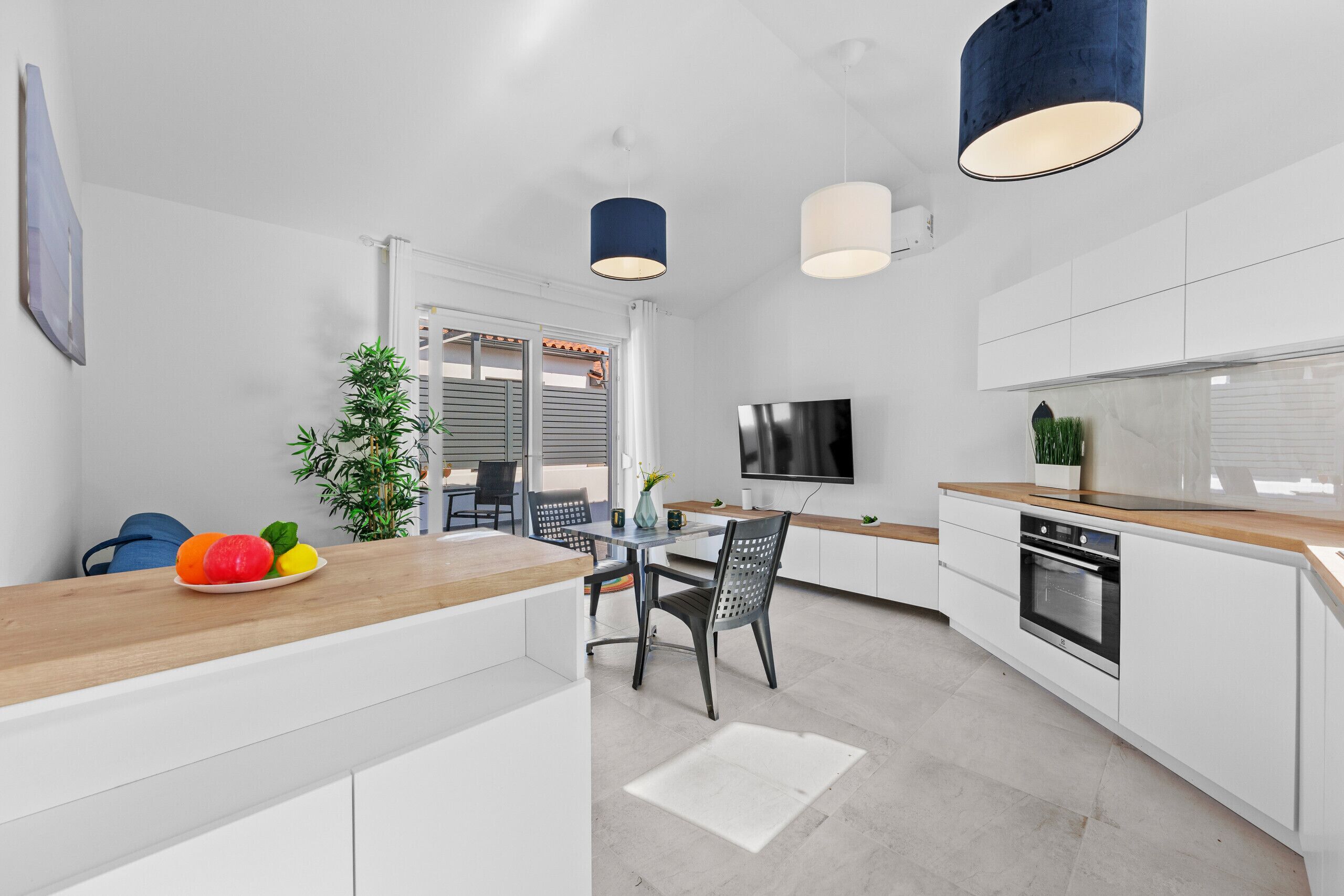 A modern kitchen with white cabinets, a wooden countertop, a dining area with chairs, a TV mounted on the wall, and a small potted plant.