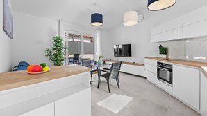 Una cocina moderna con armarios blancos, encimera de madera, zona de comedor con sillas, televisor montado en la pared y una pequeña planta en maceta.