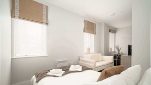 A modern hotel room with a large bed, a sofa, a chair, a TV, and a window with blinds. - Modern Studio Apartment in Waterloo, Liverpool (Liverpool)