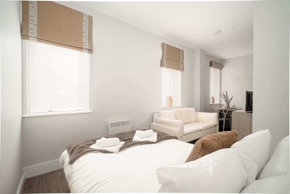 A modern hotel room with a large bed, a sofa, a chair, a TV, and a window with blinds. - Modern Studio Apartment in Waterloo, Liverpool (Liverpool)