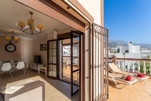 A balcony with a view of mountains, a dining area with a TV, and a living room with a chandelier. - Villa Miguel Franco (Nerja)