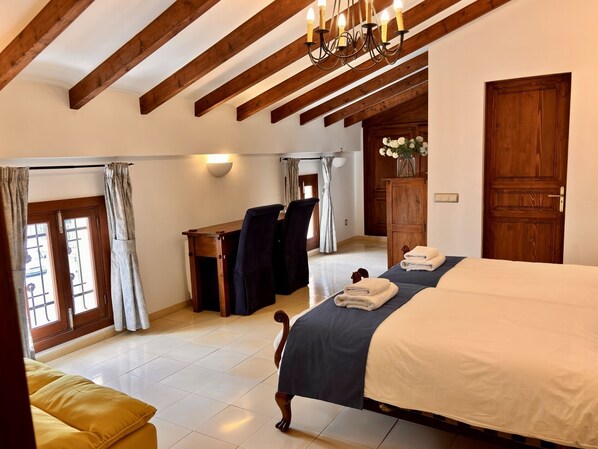 A bedroom with a bed, a desk, and a chair. There is a wooden door, a window with curtains, and a chandelier.