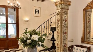 A grand foyer with a checkered floor, a round wooden table with a vase of flowers, an ornate column, a staircase with a black railing, and a large mirror.