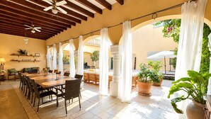 A covered outdoor dining area with a wooden table and chairs, white curtains, and potted plants.