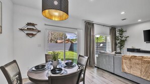 Een moderne eetruimte met een ronde tafel, stoelen en een hanglamp. Achtergrond heeft een bank en een televisie.