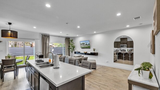 Een moderne keuken met een centraal eiland, een eethoek met tafel en stoelen, een woonkamer met een bank, en uitzicht op de achtertuin door grote ramen.
