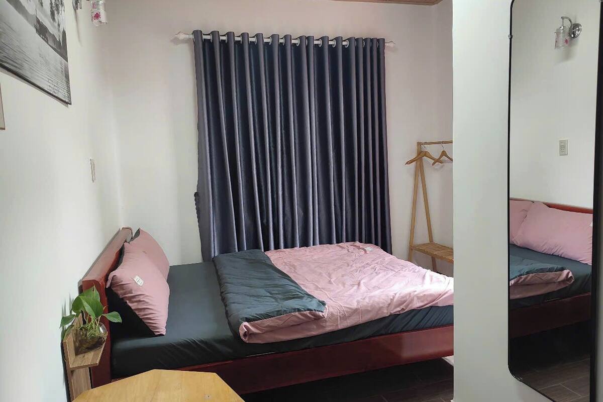 Une chambre avec un lit simple, une table de chevet en bois, un miroir et une fenêtre avec des rideaux foncés.