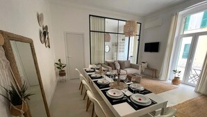 A dining area with a long table set for six, white chairs, a mirror, a television, and a view of the outdoors through a sliding door.