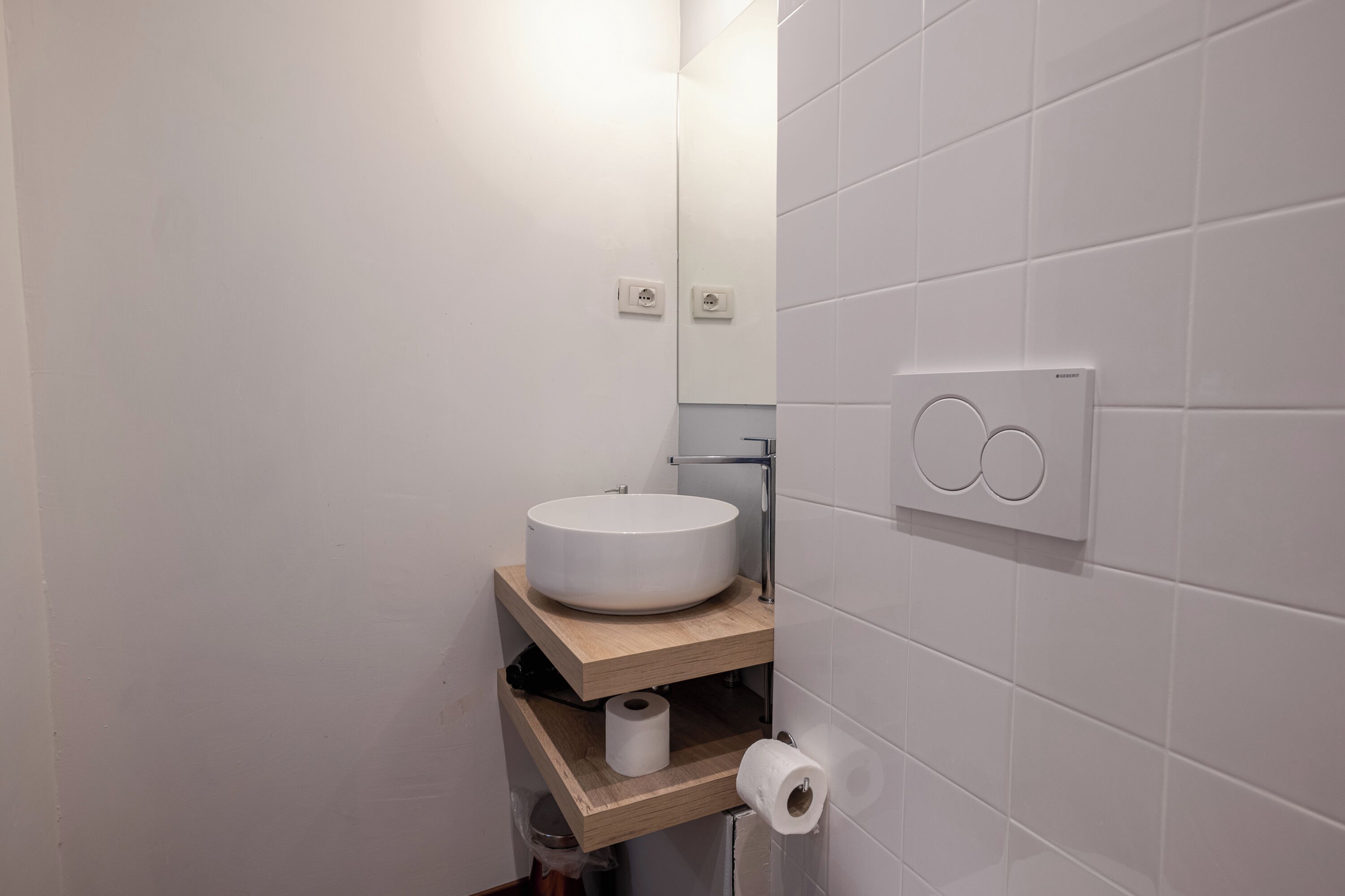 Un baño moderno con un lavamanos blanco y redondo, estantes de madera, un rollo de papel higiénico y un interruptor de luz en la pared.