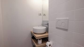 Un baño moderno con un lavamanos blanco y redondo, estantes de madera, un rollo de papel higiénico y un interruptor de luz en la pared.