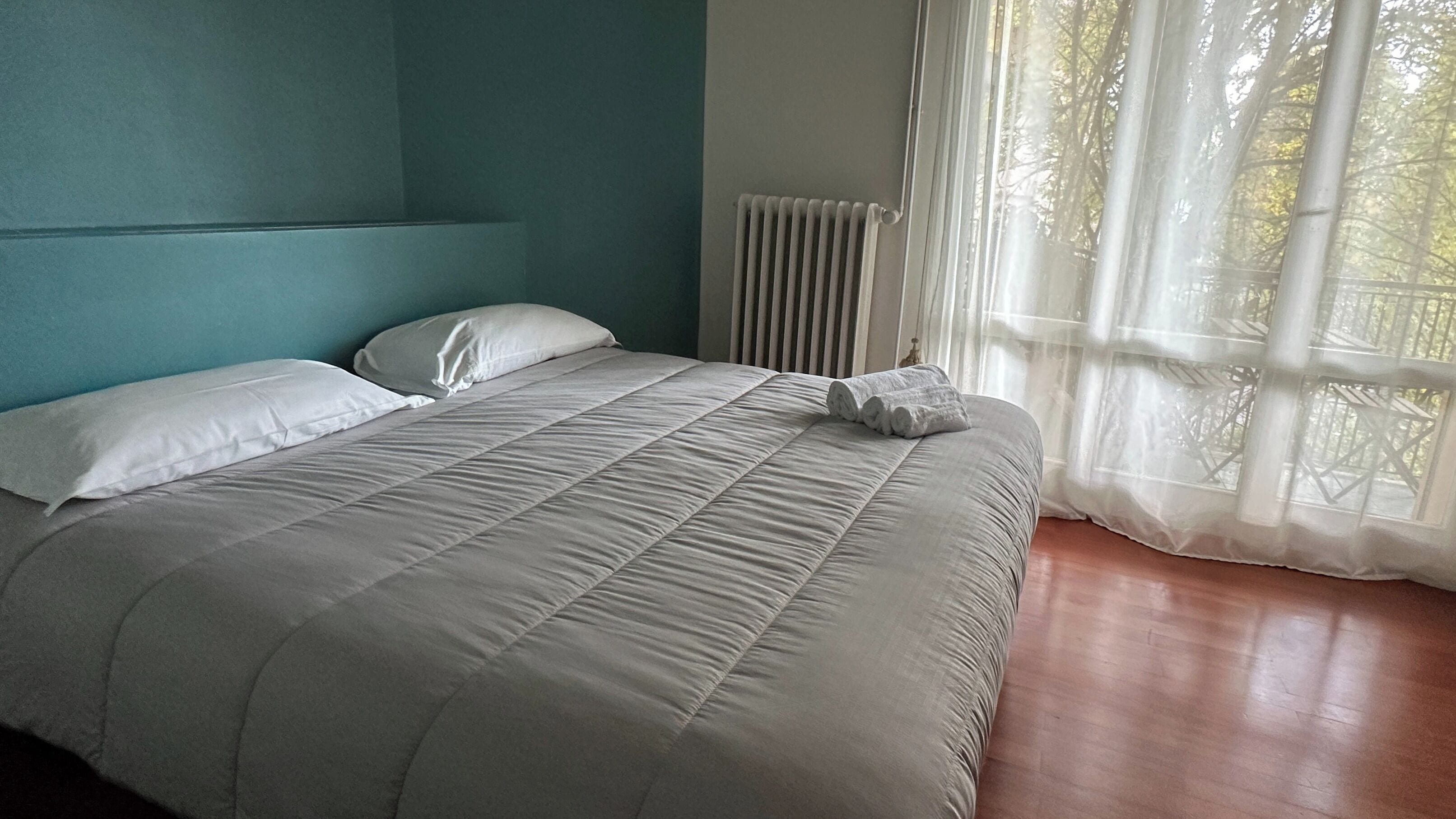 Ein Schlafzimmer mit einem Bett, zwei Kissen, einem Heizkörper und einem Fenster mit transparenten Vorhängen.