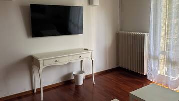 Une pièce avec un bureau blanc, un téléviseur fixé au mur, un radiateur et une fenêtre ornée de rideaux légers.