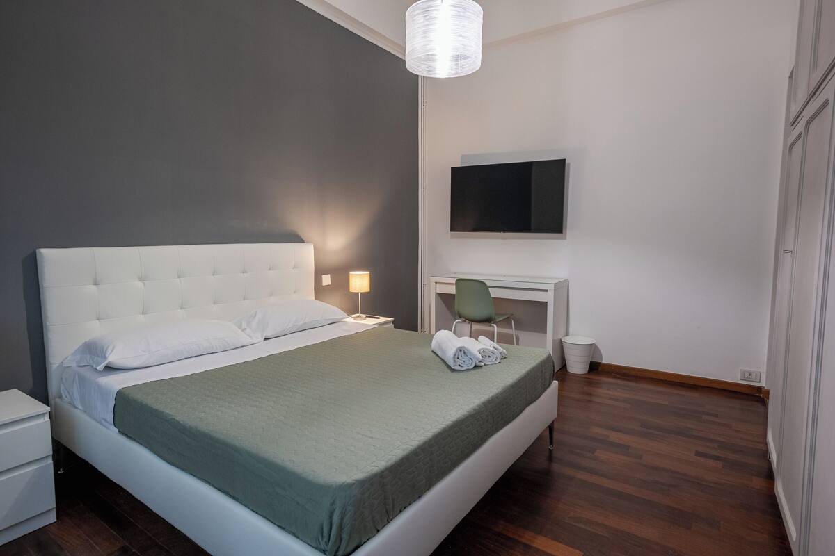 A modern bedroom with a large bed, a desk, and a TV mounted on the wall.