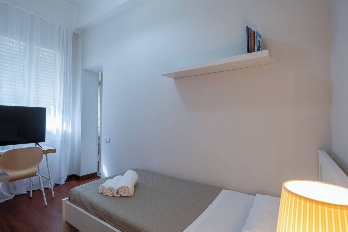 A hotel room with a bed, a desk with a chair, a television, and a shelf with books.