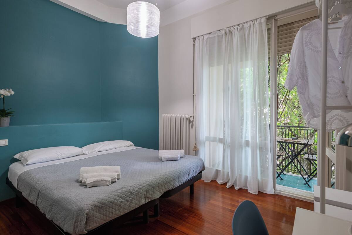 A bedroom with a bed, a radiator, a window with curtains, and a balcony with a table and chairs.