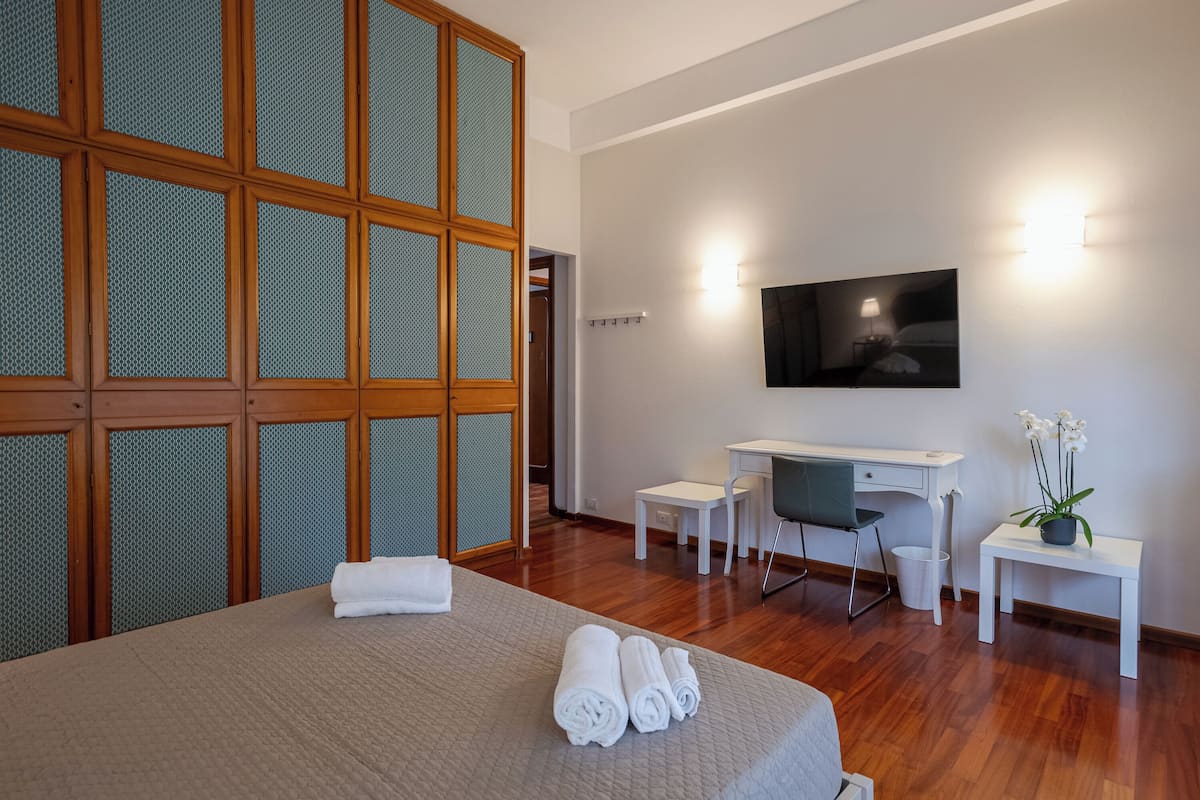 A hotel room with a bed, a desk, a chair, a TV, and a small table with a plant.