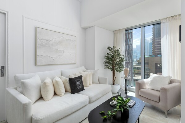 A modern living room with a white sofa, a black coffee table, and a beige armchair. There is a large window with a city view, a potted plant, and a framed artwork on the wall.