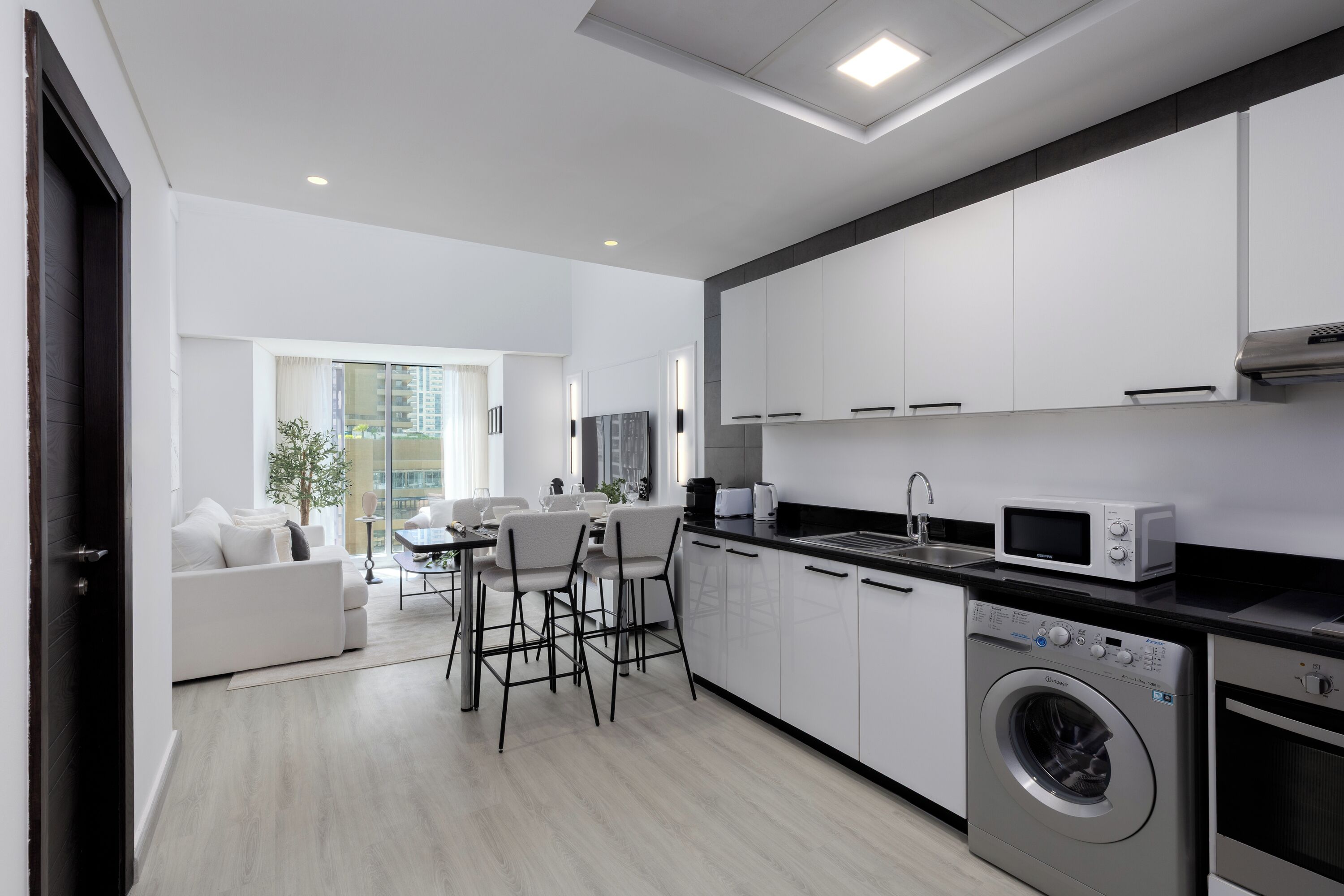 Eine moderne Küche mit weißen Schränken, einer integrierten Waschmaschine und einem Trockner, einem Essbereich mit weißem Sofa und Stühlen sowie einem großen Fenster, das für natürliches Licht sorgt.