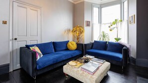 A living room with two blue sofas, a coffee table with books, a yellow cushion, and a large plant.