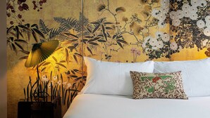 A bed with white linens and a patterned pillow, a wall with a floral mural, and a lamp with a yellow shade.