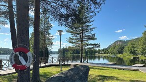 Un parc au bord du lac, équipé d’une bouée de sauvetage, de bancs en bois, d’un grand rocher et offrant une vue dégagée sur le lac et les collines environnantes.