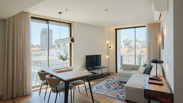 Ein modernes Wohnzimmer mit Essbereich, einer Couch und einem Fernseher. Große Fenster bieten Blick auf die Stadt.